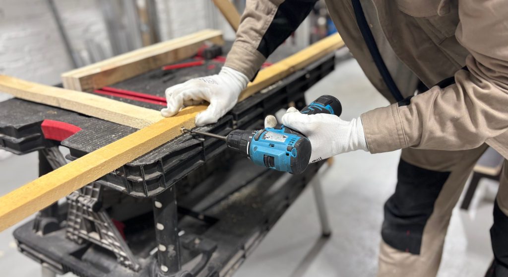 A professional tradesperson wearing safety gloves and using a cordless drill to drive a screw into timber on a workbench.